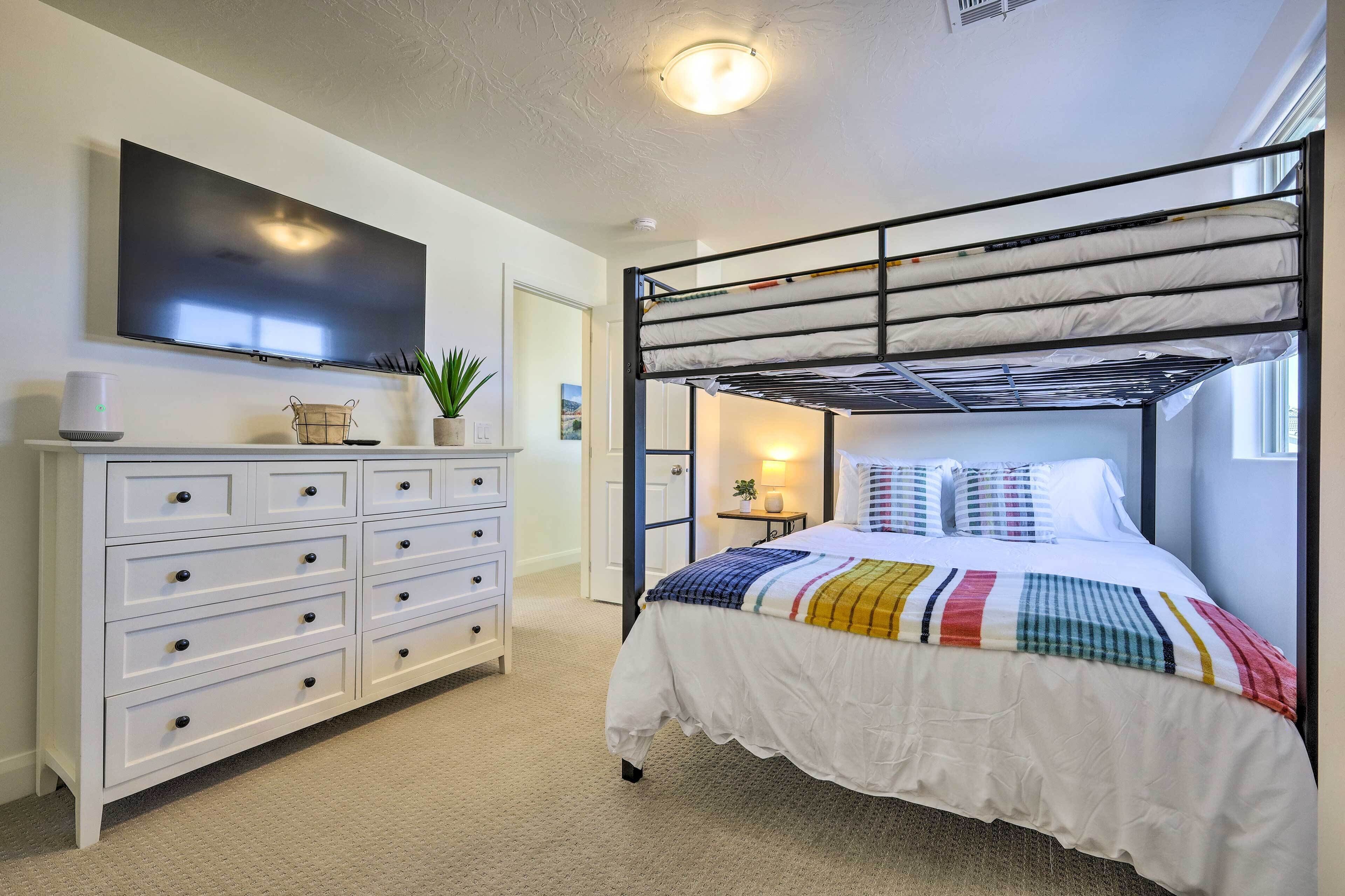 A bedroom with a bunk bed, a TV mounted on the wall, a white dresser with drawers, and a bedside table with a lamp.