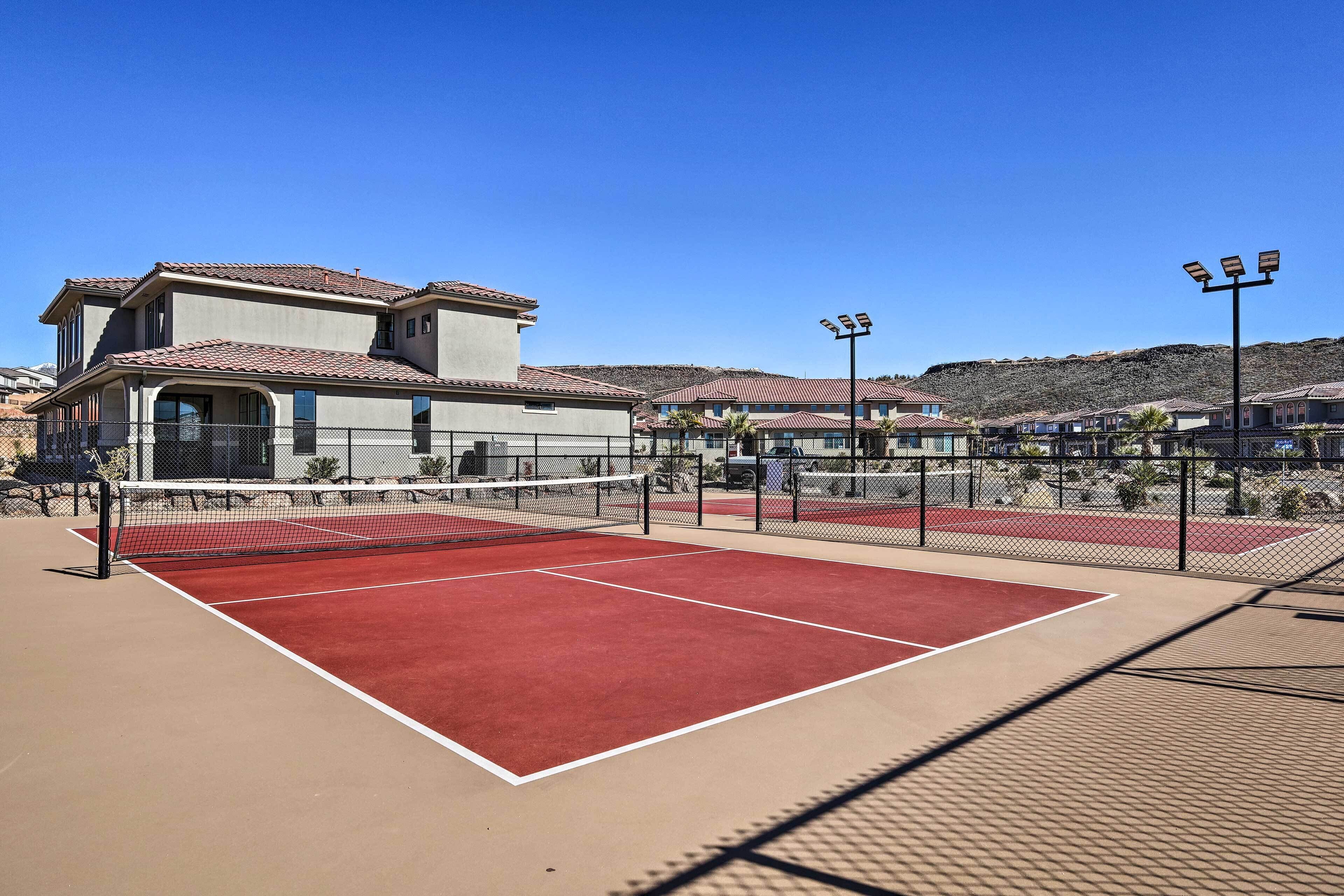 Un court de tennis extérieur, doté d’un filet et entouré d’une clôture, jouxtant un bâtiment dont le toit est recouvert de tuiles rouges.
