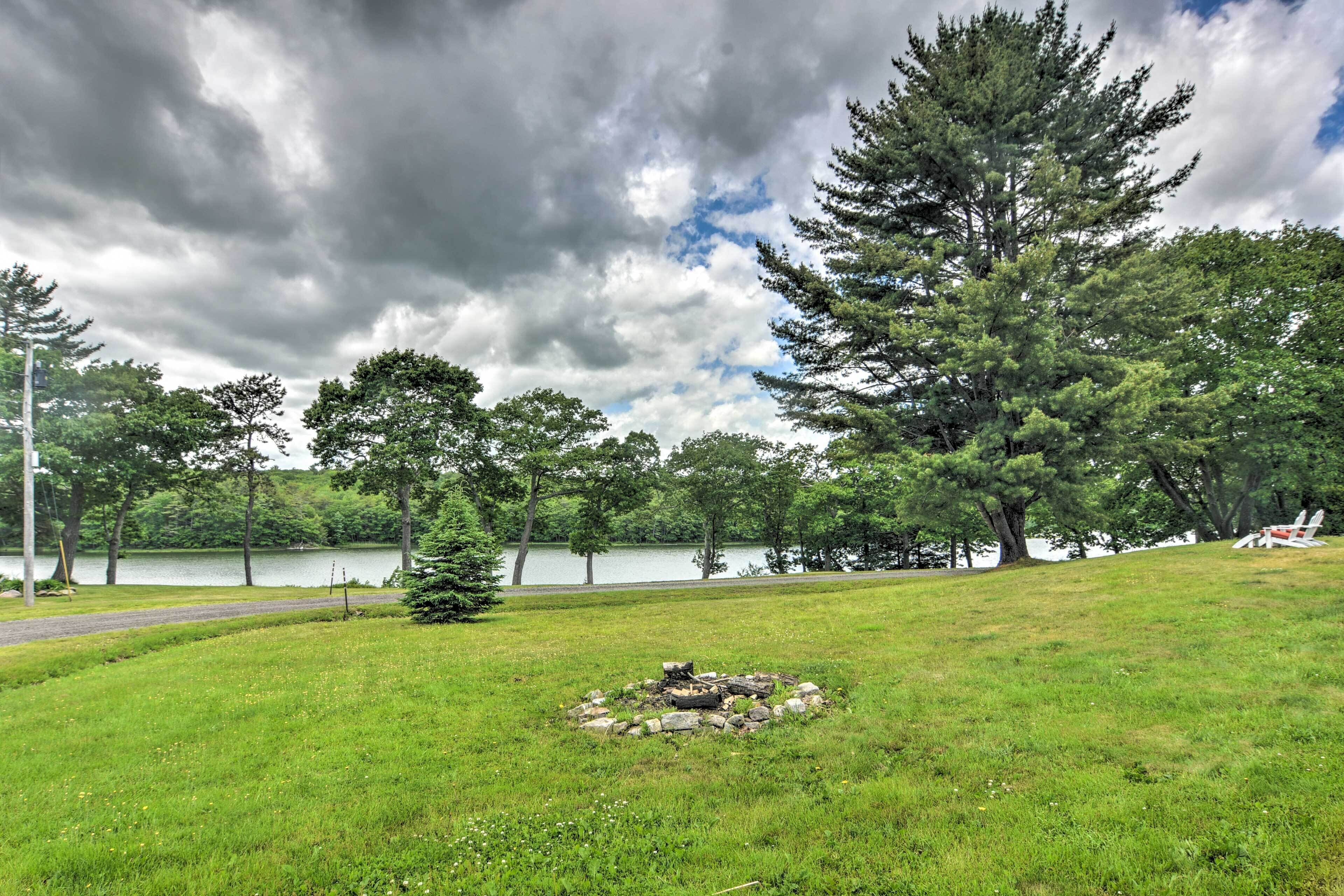 A grassy area with a fire pit, trees, and a lake in the background.