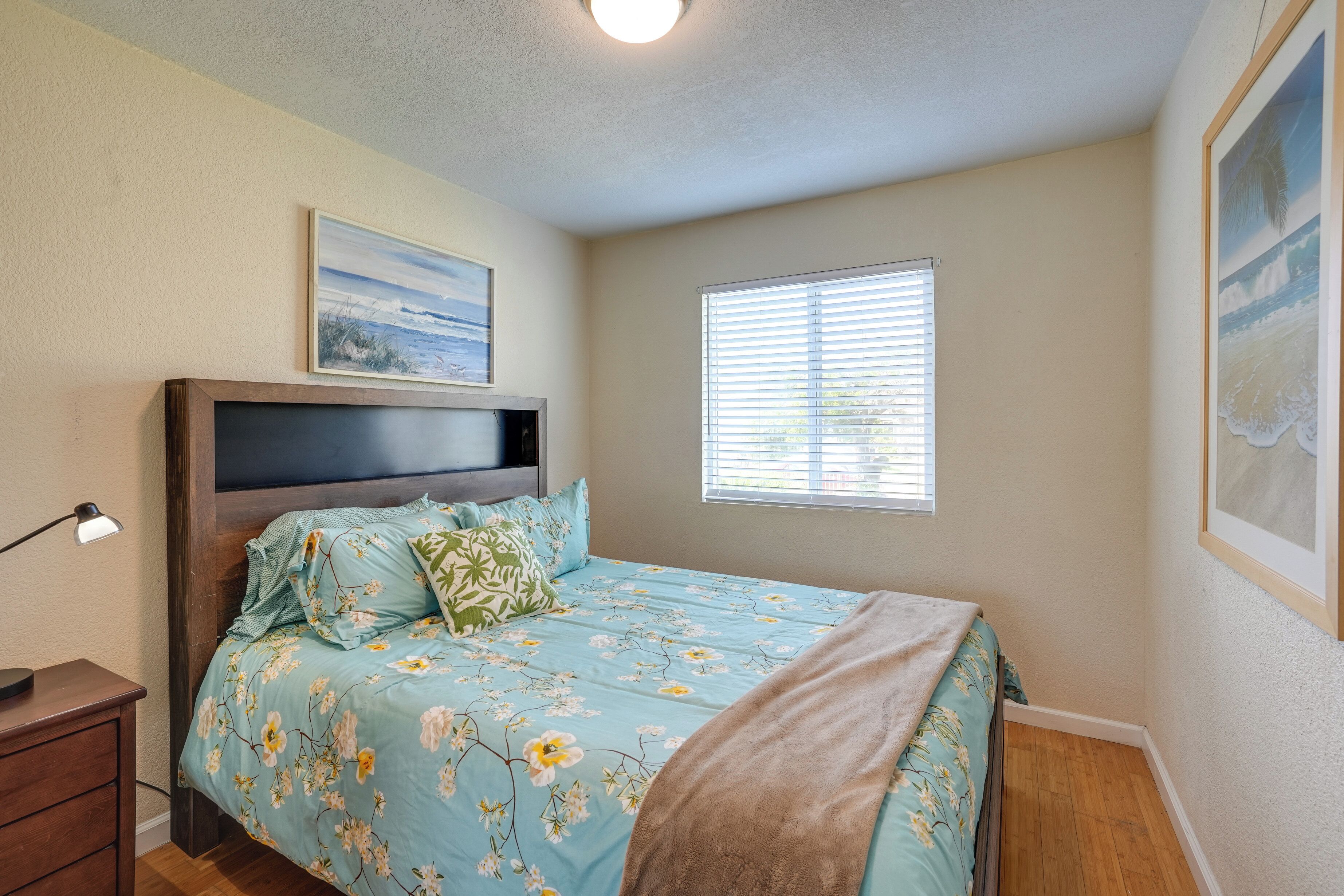 A bedroom with a bed, a nightstand, a window with blinds, and a painting on the wall.