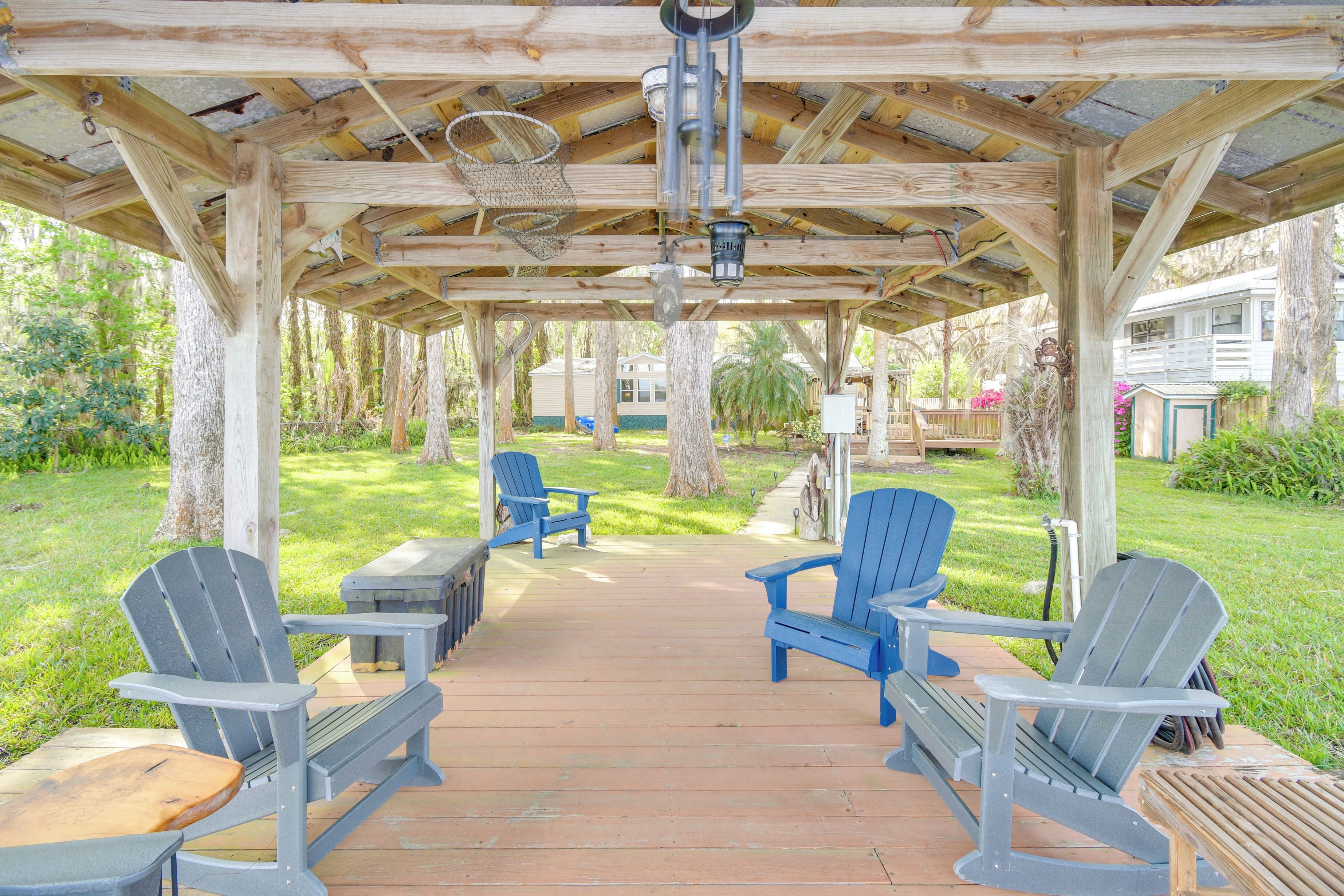 A wooden deck with Adirondack chairs and a table, surrounded by greenery and a covered area with a basketball hoop.
