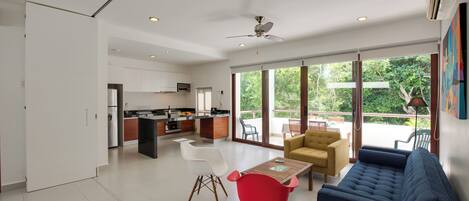 A modern kitchen with white cabinets, a dining area with a red chair and a blue sofa, and a balcony with a view of greenery.