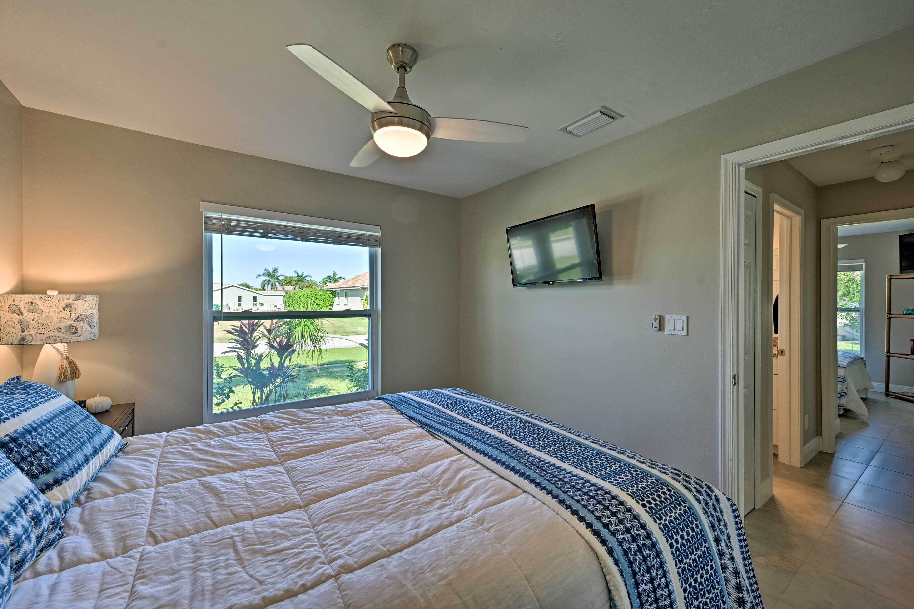 A bedroom with a bed, a ceiling fan, a TV mounted on the wall, and a window with a view of the outdoors.