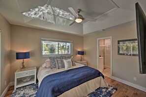 Un dormitorio con cama, mesitas de noche, ventilador de techo, ventana y puerta que da a un baño.