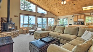 A cozy living room with a stone fireplace, large windows, a sectional sofa, and a kitchen area. - Near Hiking: Home w/ Deck in Twain Harte! (Mi-Wuk Village)