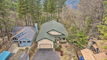 A two-story house with a large garage, surrounded by dense forest and mountains in the background.