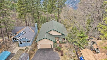 A two-story house with a large garage, surrounded by dense forest and mountains in the background.