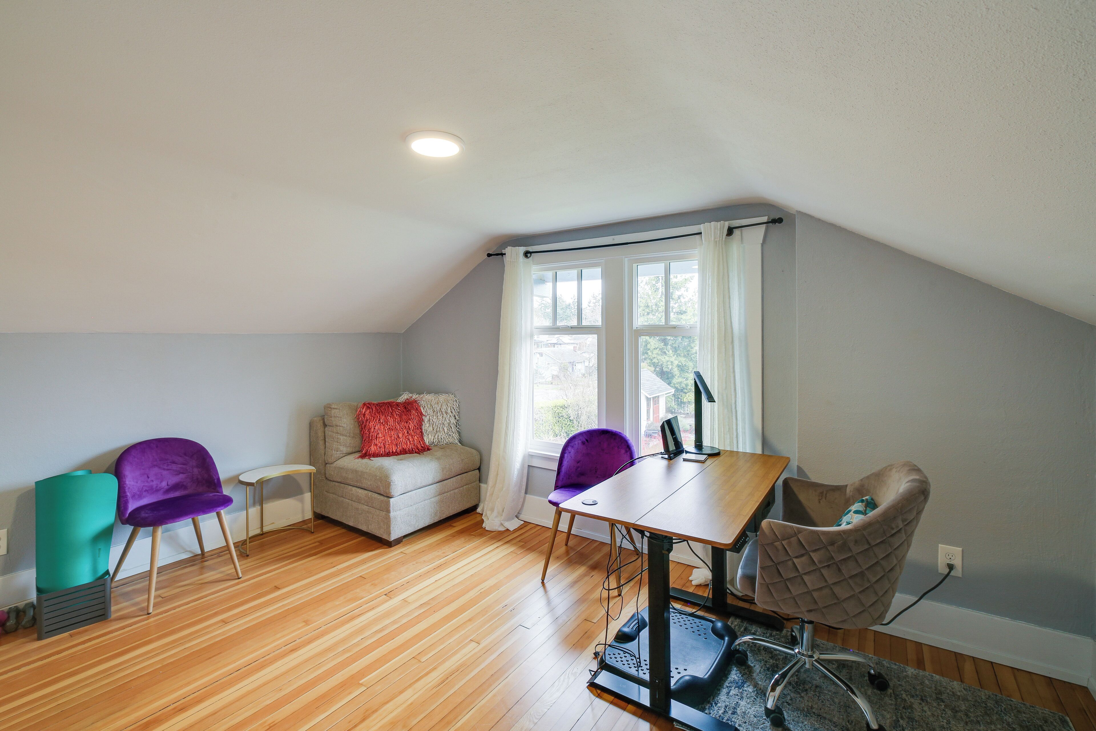 A room with a wooden floor, a sofa, a purple chair, a table with a computer, and a window with curtains.