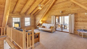 A log cabin interior with a loft bedroom, a living area with a sofa and a dining table, and a large window providing natural light. - Cottonwood Cabin w/ Private On-site Fly Fishing! (Blanco)