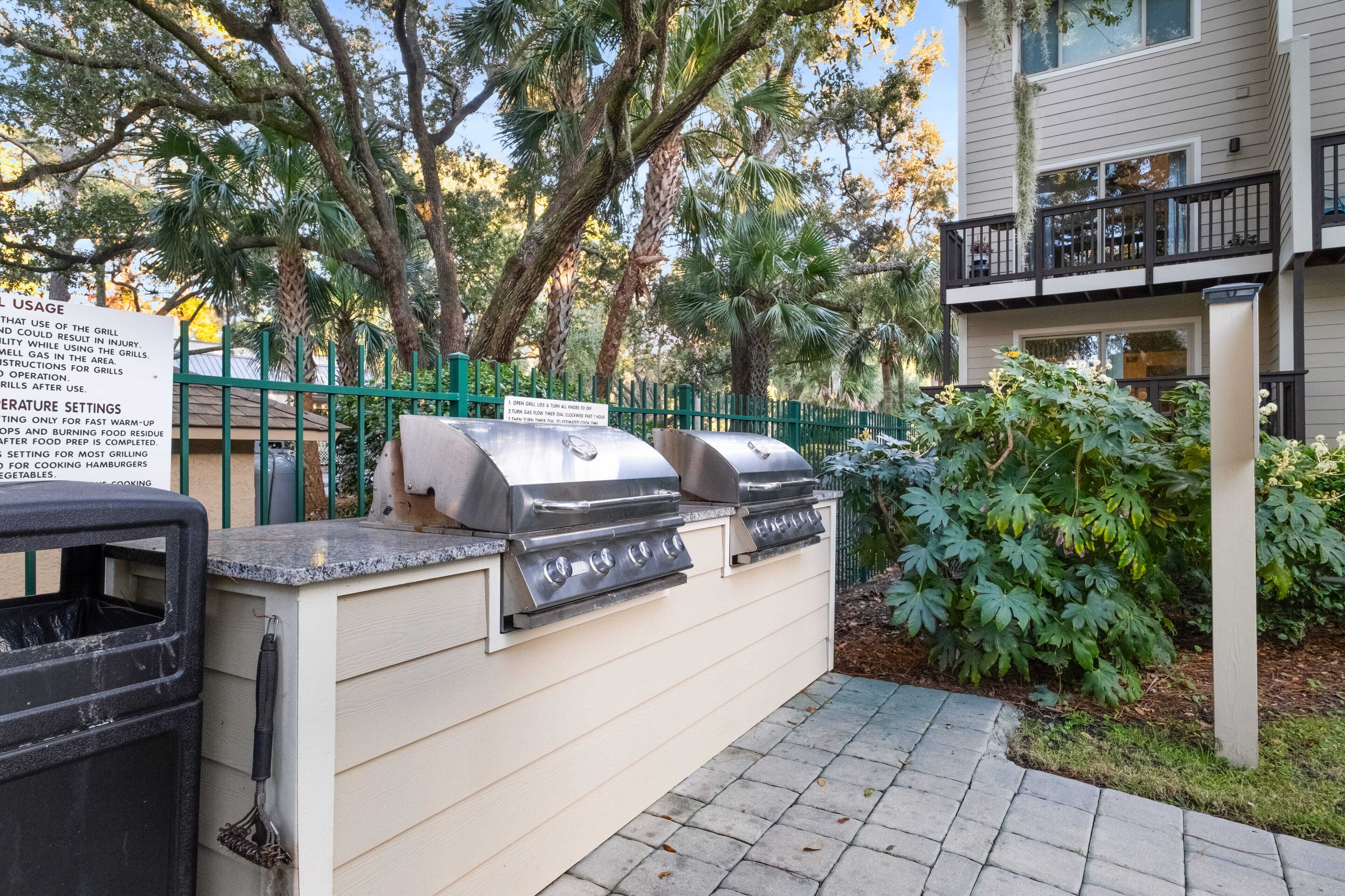 An outdoor grill area with a sign detailing usage and temperature settings, surrounded by lush greenery and a building in the background.