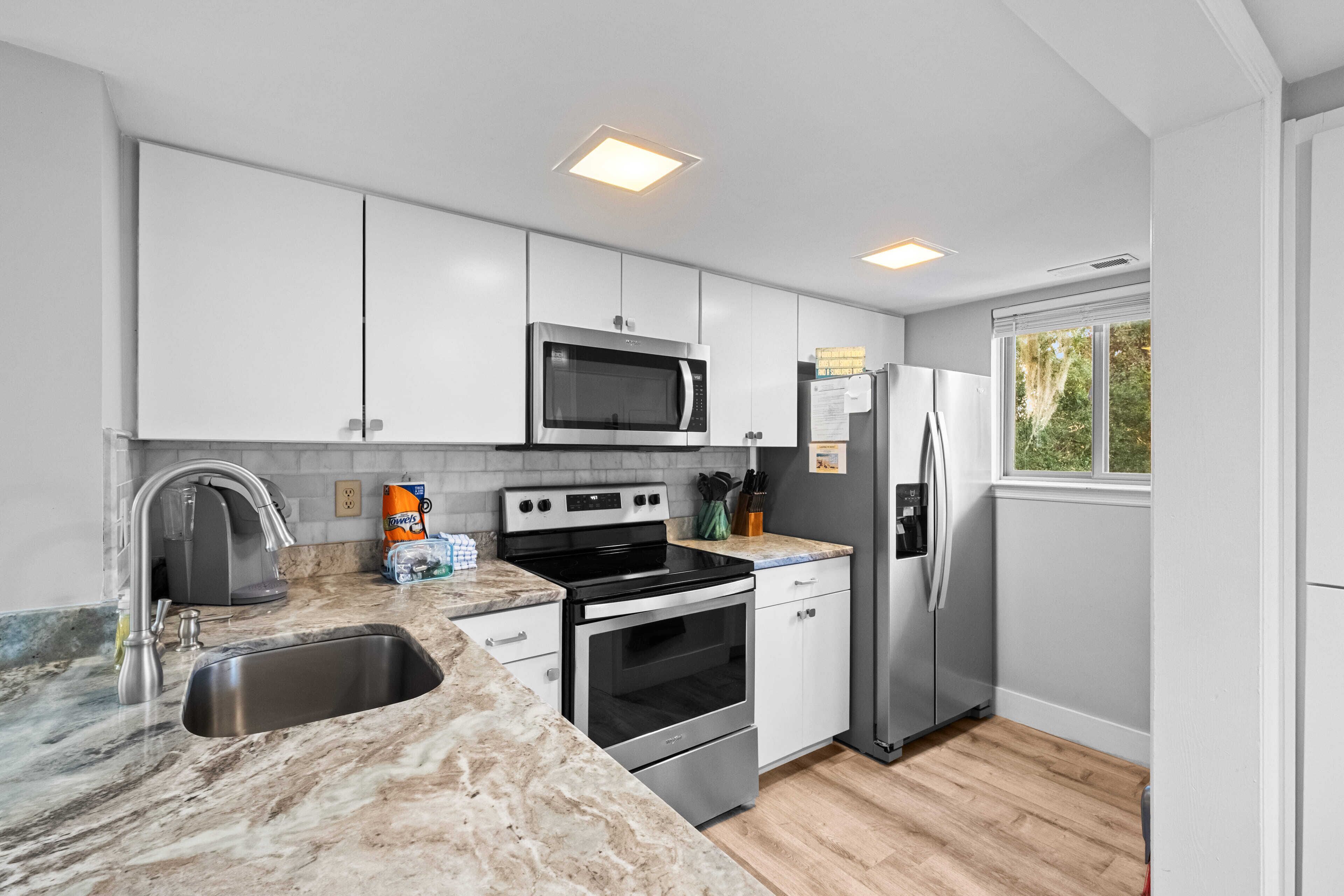 A modern kitchen with white cabinets, stainless steel appliances, a marble countertop, and a window providing natural light.