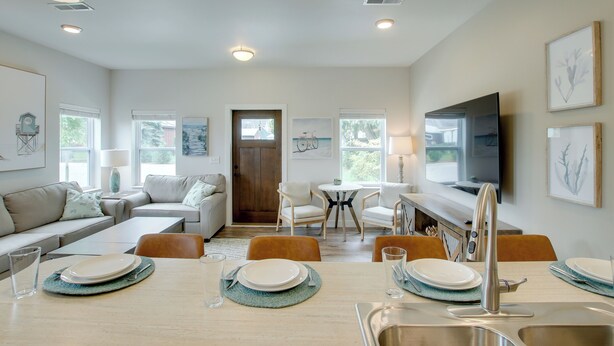 A modern living room with a dining area, featuring a sofa, chairs, a table set for dining, a TV, and artwork on the walls. - Riverfront Sheboygan Townhome w/ Grill! (Sheboygan)