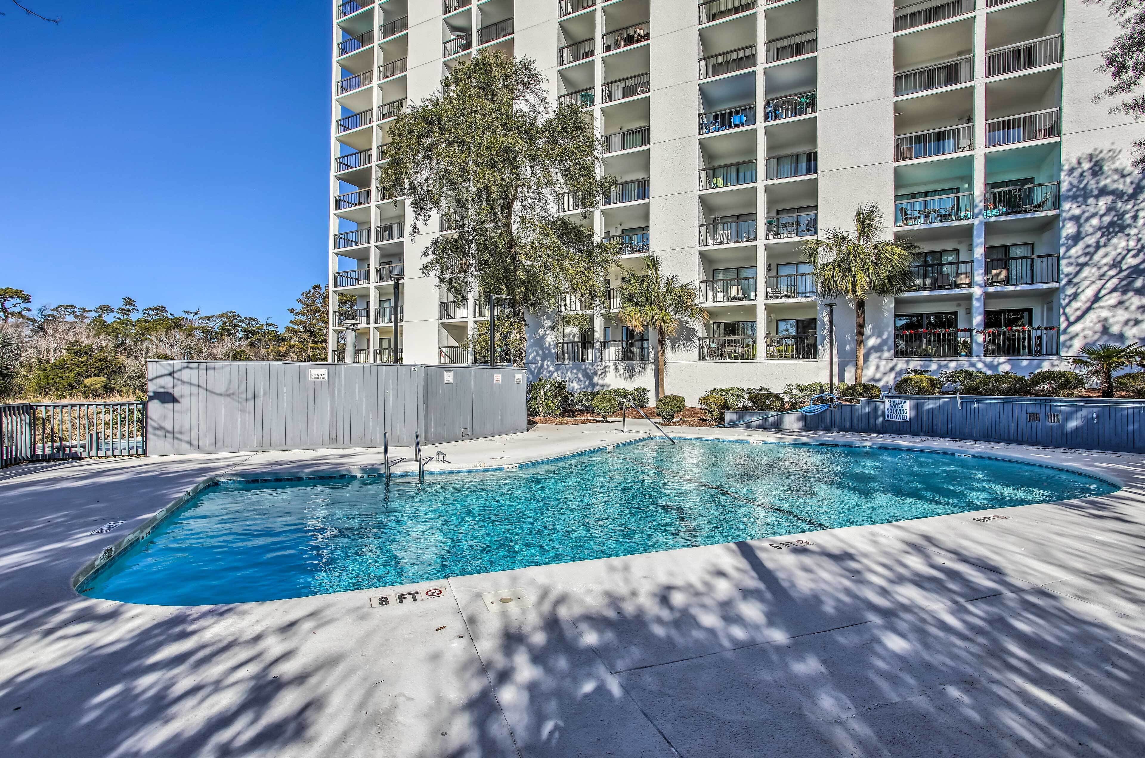 A swimming pool with a clear blue water, surrounded by a concrete deck, and adjacent to a multi-story building with balconies.