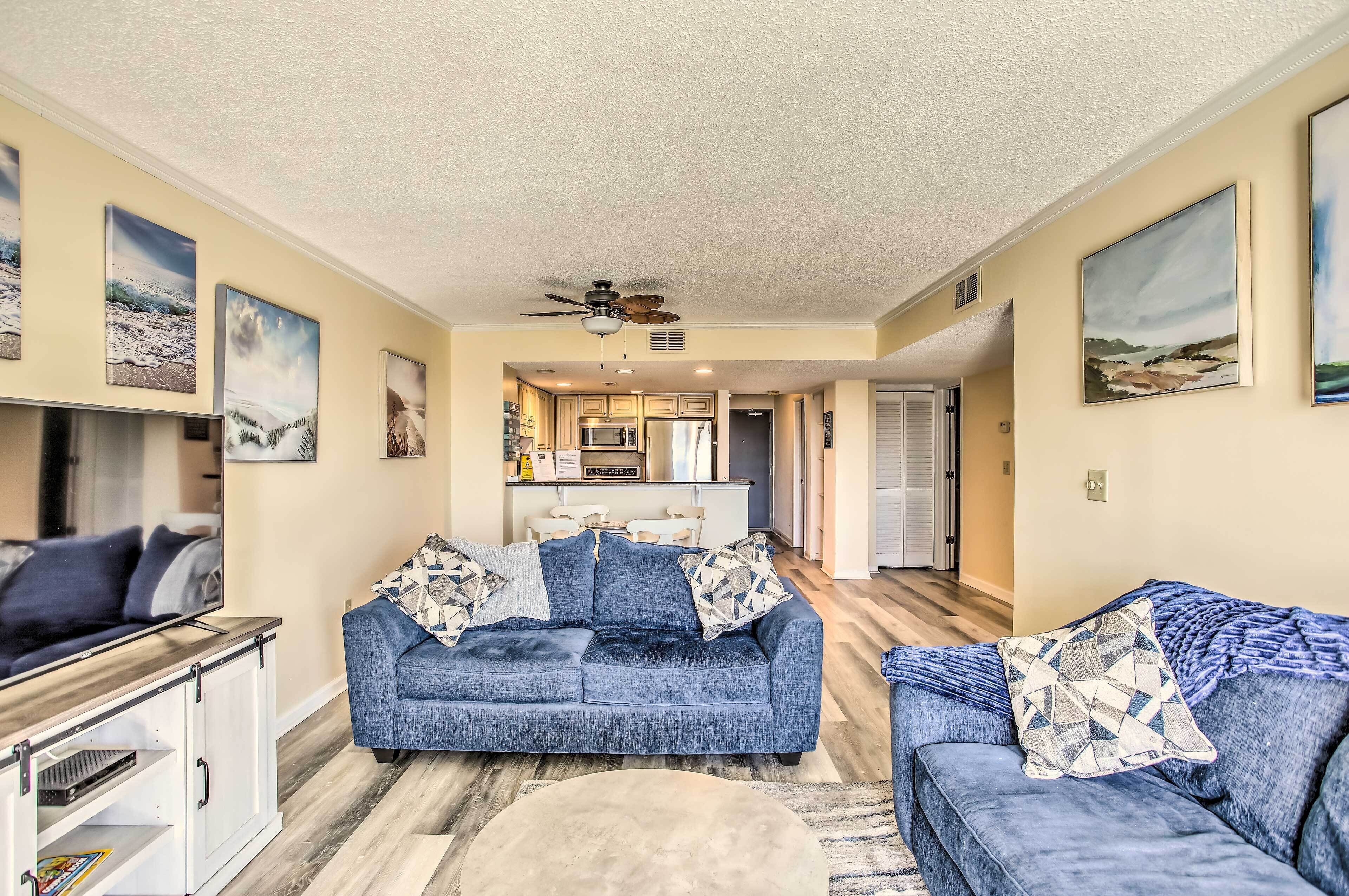 A living room with a blue sofa, a coffee table, and a TV stand. There is a ceiling fan and artwork on the walls.