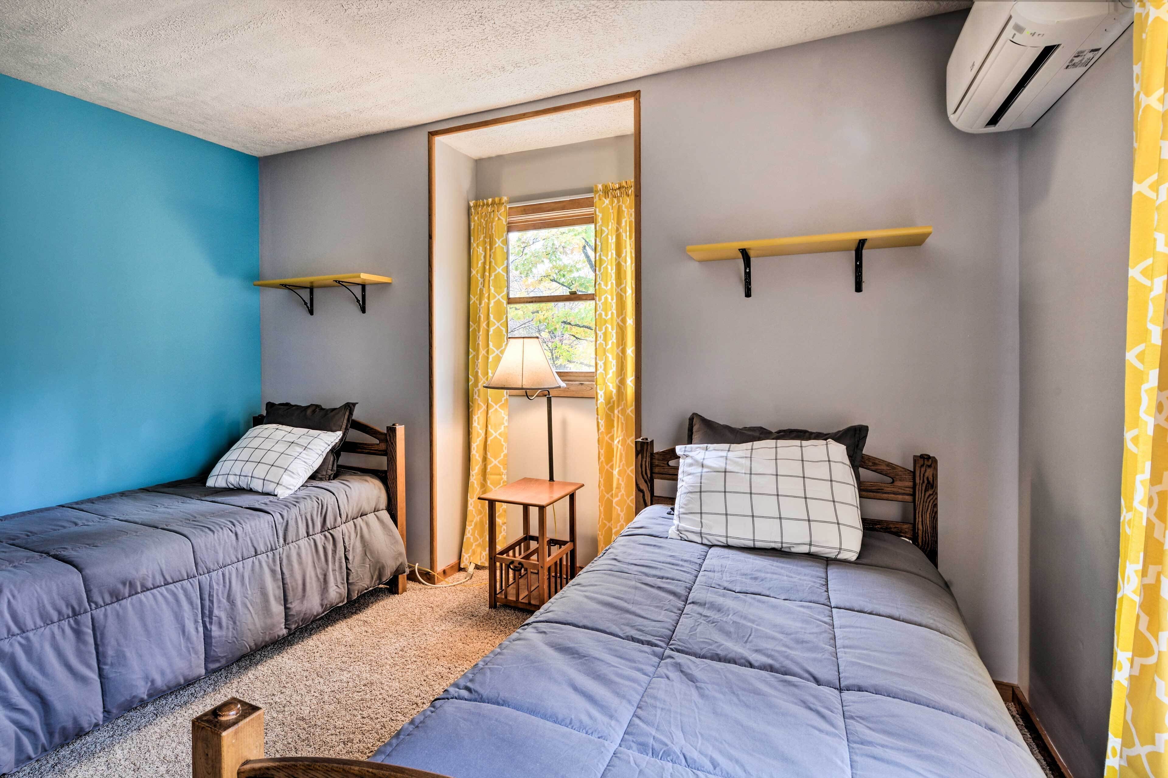 Ein Schlafzimmer mit zwei Betten, einem Fenster mit gelben Vorhängen, einem Nachttisch und einem an der Wand befestigten Regal.