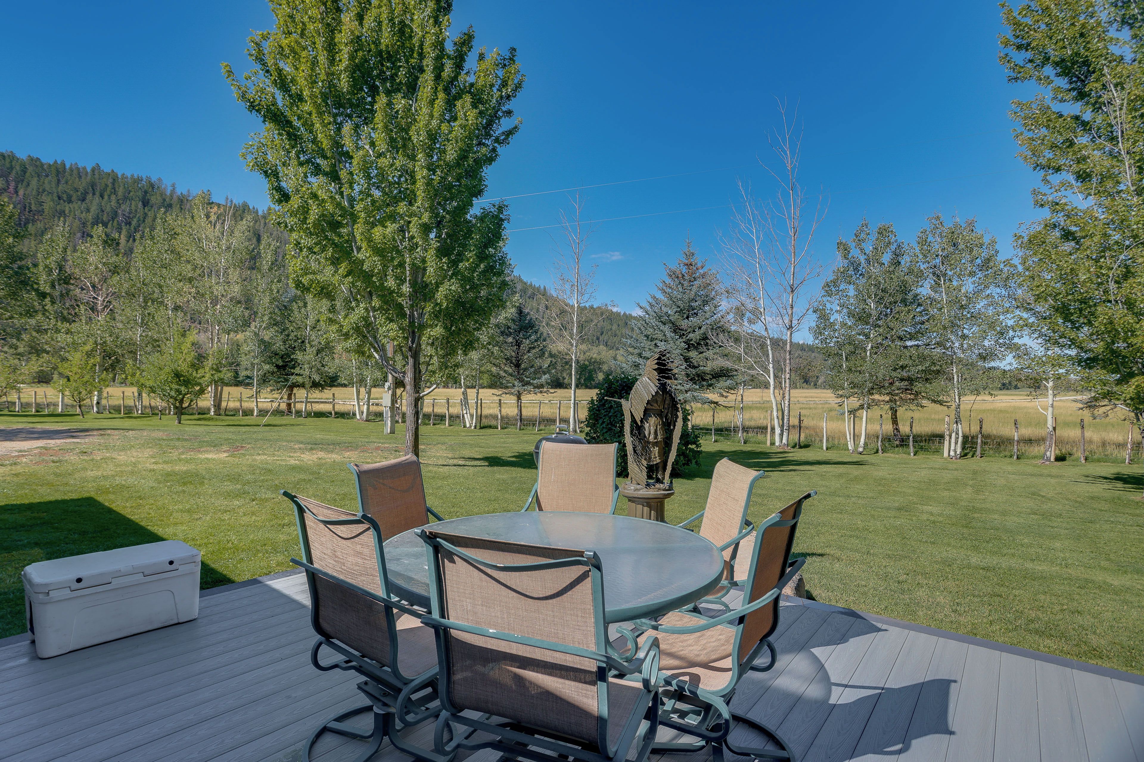 An outdoor deck with a round table and chairs, surrounded by a grassy area with trees and a fence.