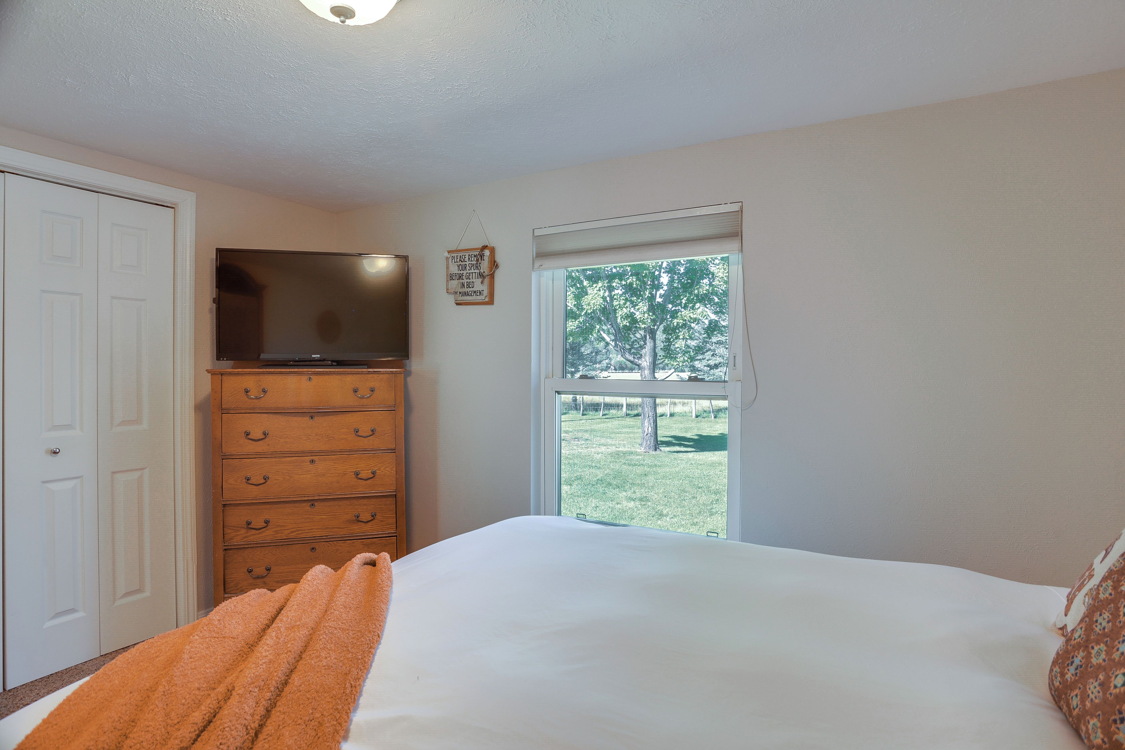 Un dormitorio con cama, cómoda, televisor y ventana con vista a árboles.