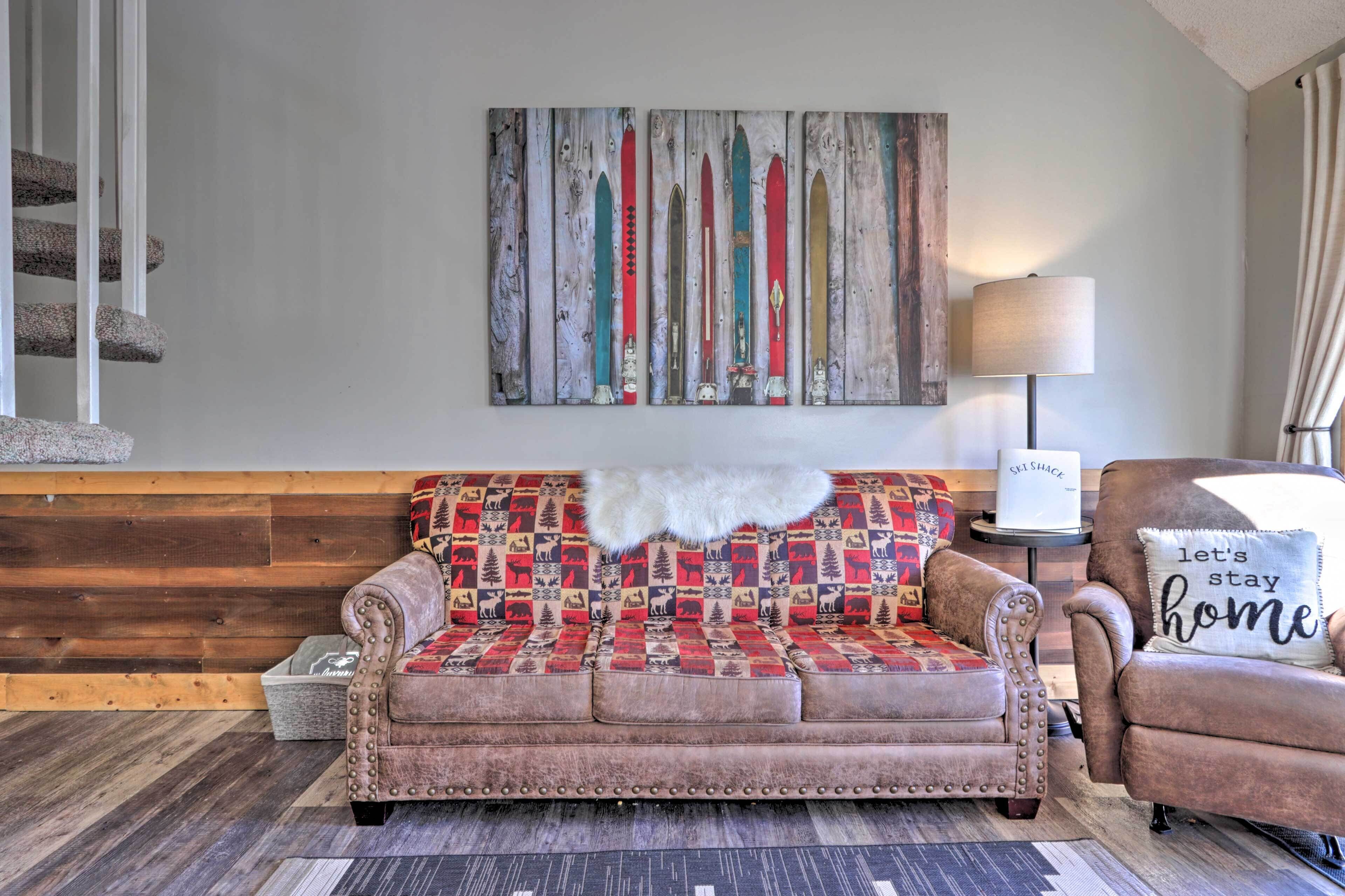 A cozy living room with a patterned sofa, a decorative wall piece, a lamp, and a 'let's stay home' pillow.