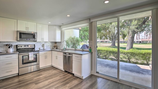 Una cocina moderna con gabinetes blancos, electrodomésticos de acero inoxidable y un amplio ventanal corredera que da a un patio con vistas a árboles y un edificio.
