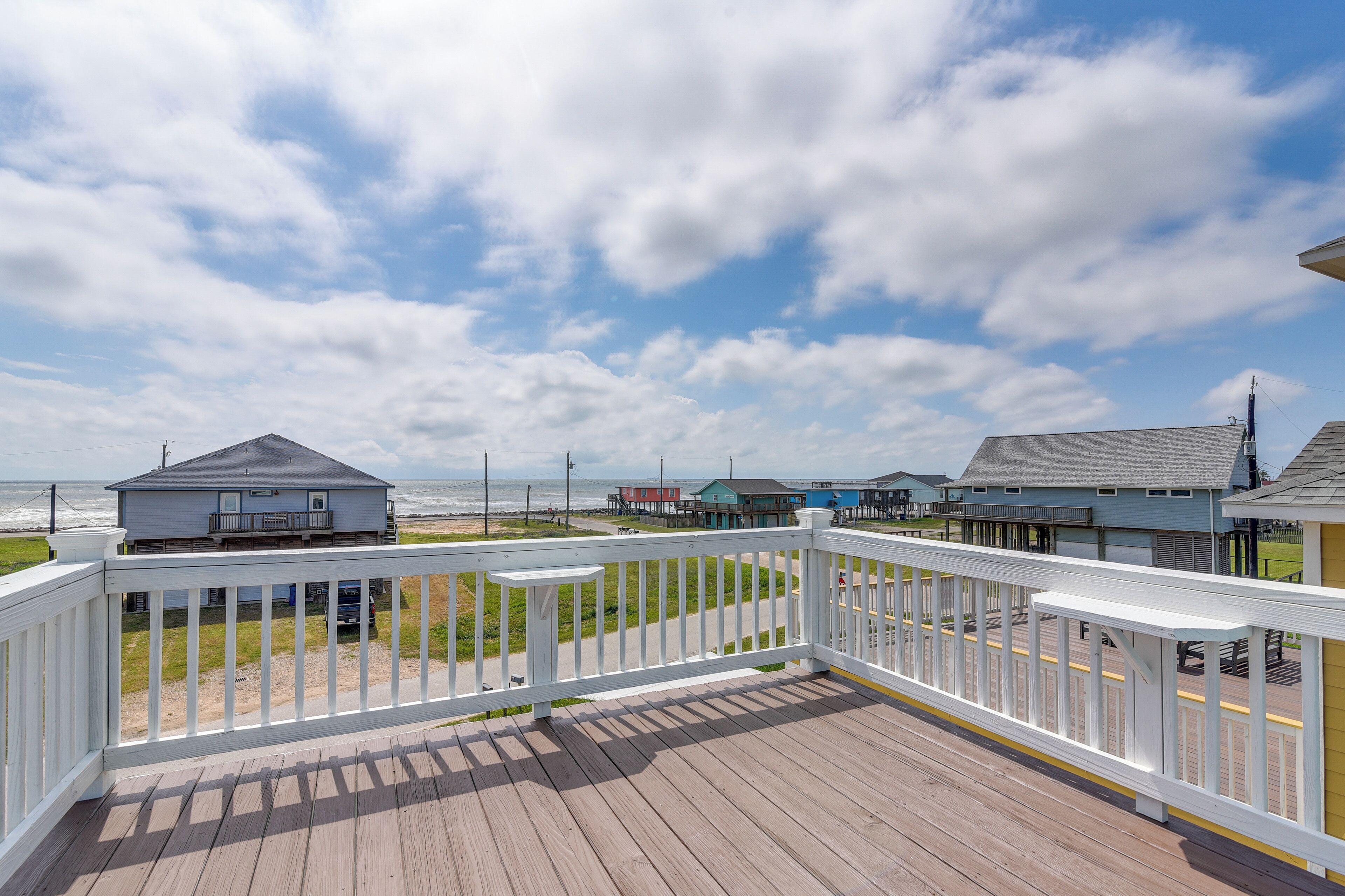 Une terrasse en bois avec des rampes blanches donnant sur une plage bordée de bâtiments.