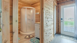 Interior de madera con cabina de ducha de vidrio, lavamanos blanco y puerta que da al exterior.