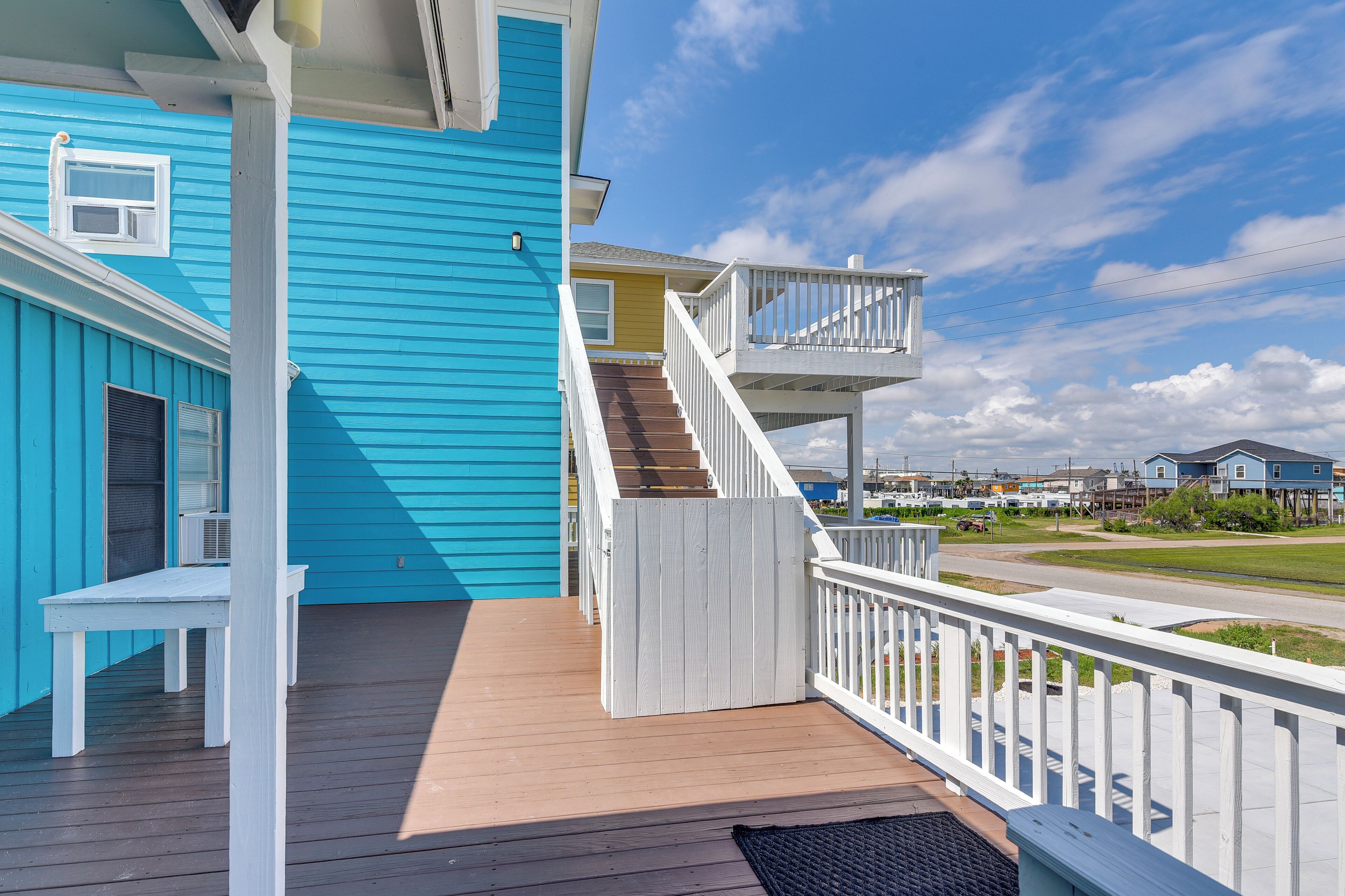 A blue building with a wooden deck, white railings, and a staircase leading to a second floor.