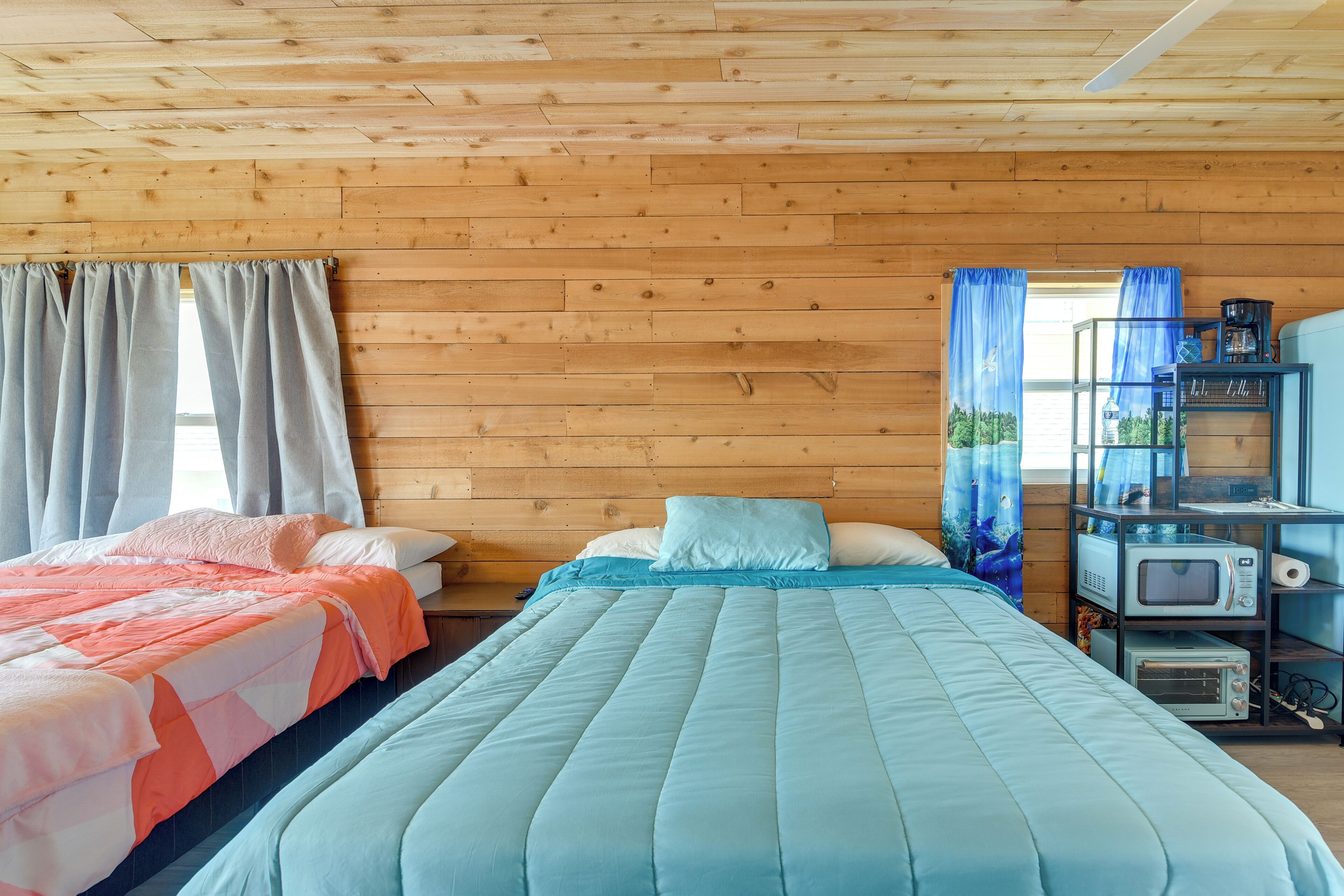 A wooden cabin room with two beds, a shelf with a microwave, and a window with curtains.
