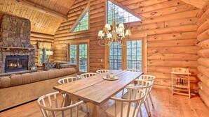 L’intérieur d’une cabane en rondins, avec une cheminée en pierre, une table à manger en bois, des chaises blanches et une grande fenêtre.
