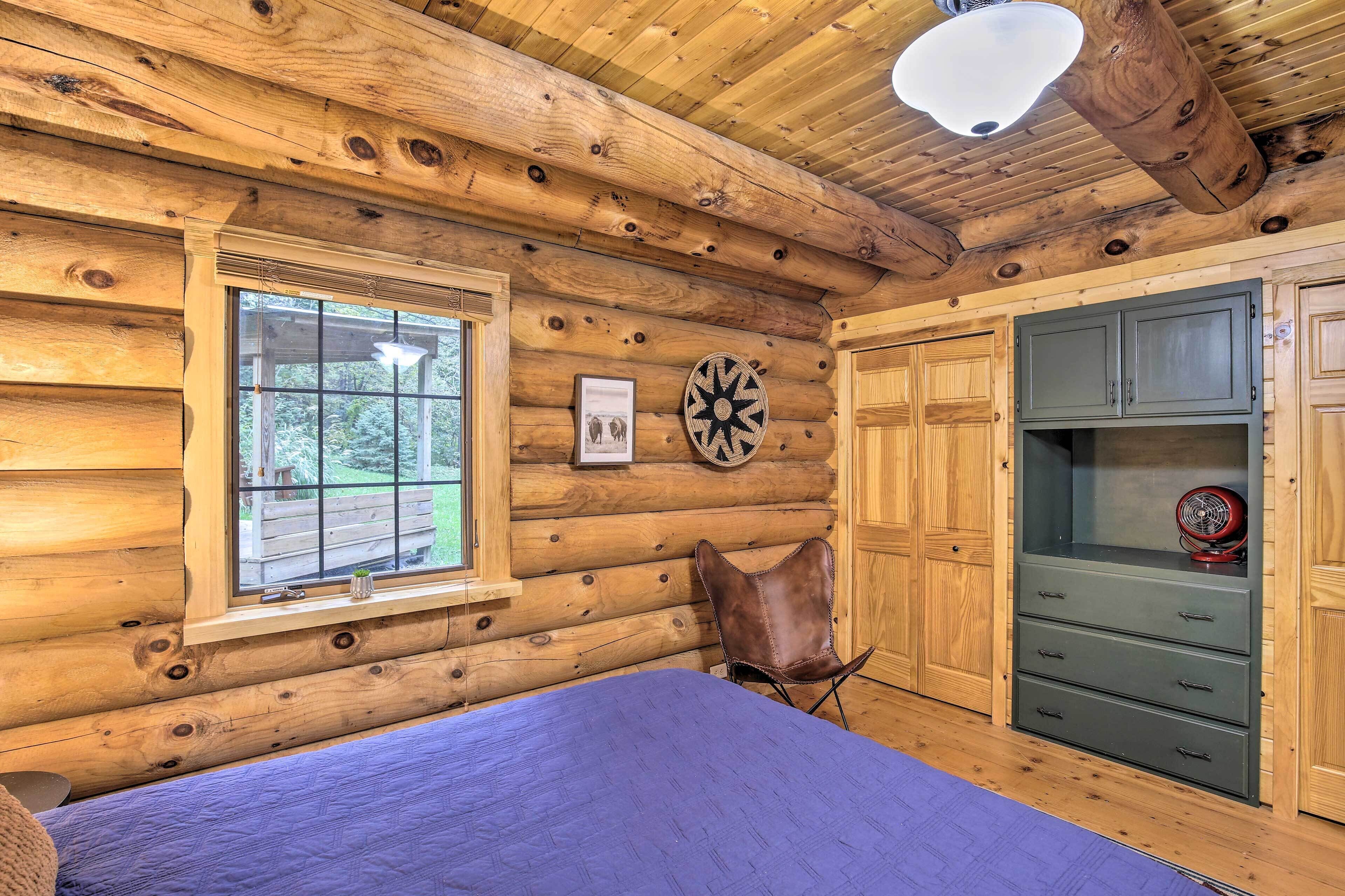 A log cabin room with a bed, a window, a wall-mounted fan, and wooden cabinets.