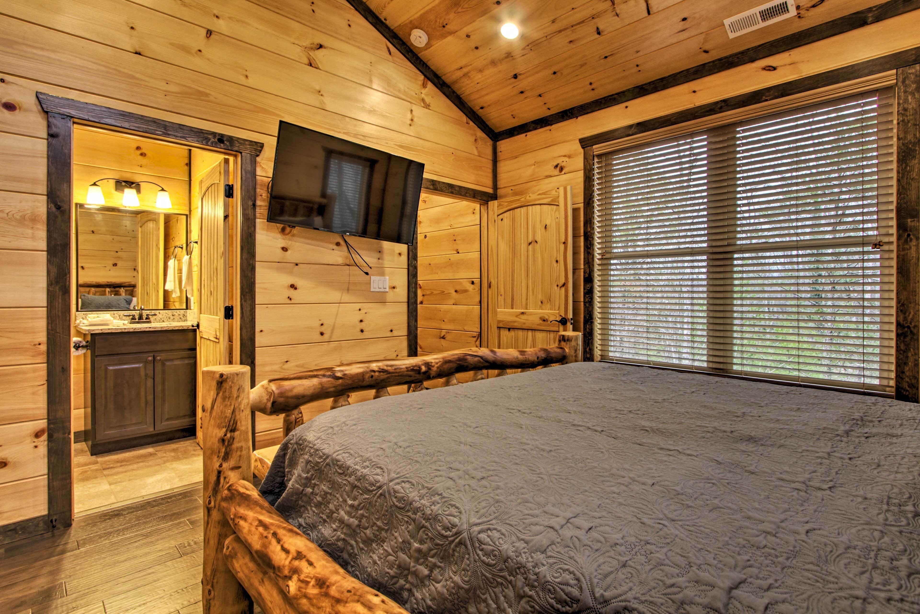 Quarto de cabana de tronco com uma cama, uma televisão na parede, um banheiro com pia e espelho, e uma janela com persianas.