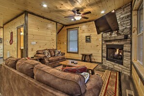 A cozy cabin interior with a stone fireplace, wooden walls, a leather sofa, and a flat-screen TV mounted on the wall. - Luxe Cabin w/ Home Theater < 2 Miles to Gatlinburg (Gatlinburg)