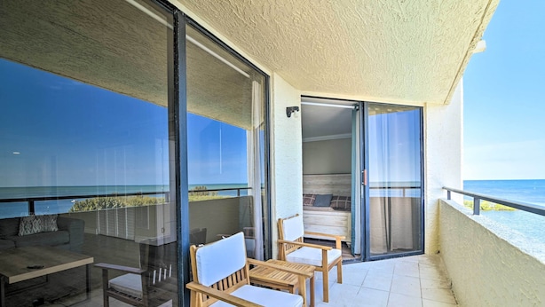 A balcony with a glass door, wooden chairs, a small table, and a sofa. - Waterfront Condo: Resort Pool, Tennis & Beach (Hudson)