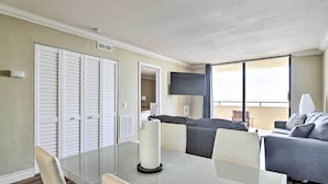 A modern living room with a glass dining table, grey sofa, and a TV mounted on the wall. - Waterfront Condo: Resort Pool, Tennis & Beach (Hudson)