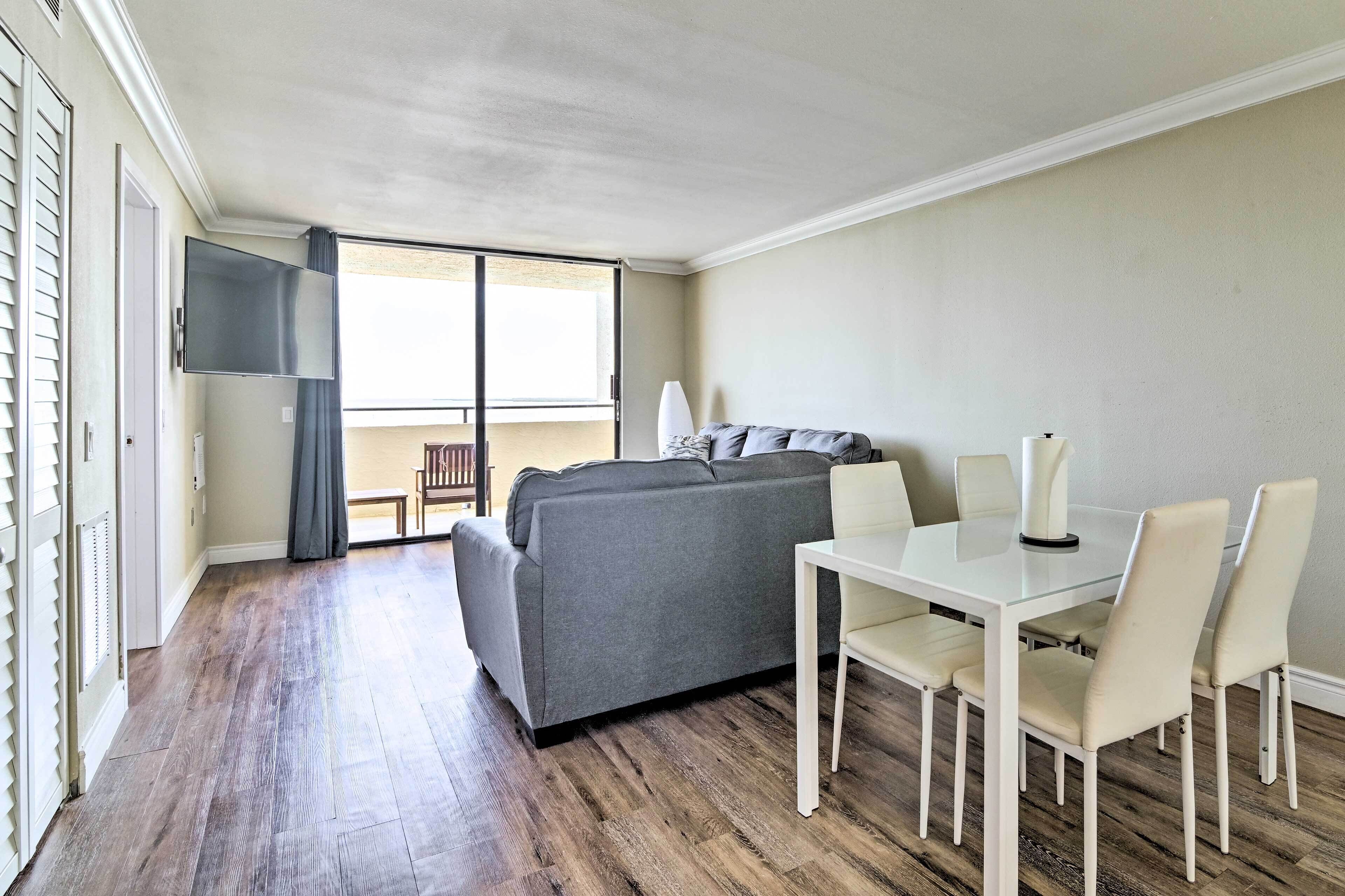 A modern living room with a grey sofa, a white dining table, and a TV mounted on the wall.