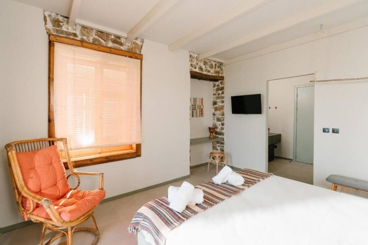 A bedroom with a bed, a wicker chair, a window with blinds, a stone wall, a television, and a bathroom door.