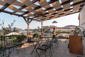 Eine Terrassengaststätte mit Holztischen und -stühlen, einer Pergola mit hängenden Lampen und Blick auf die Skyline im Hintergrund.