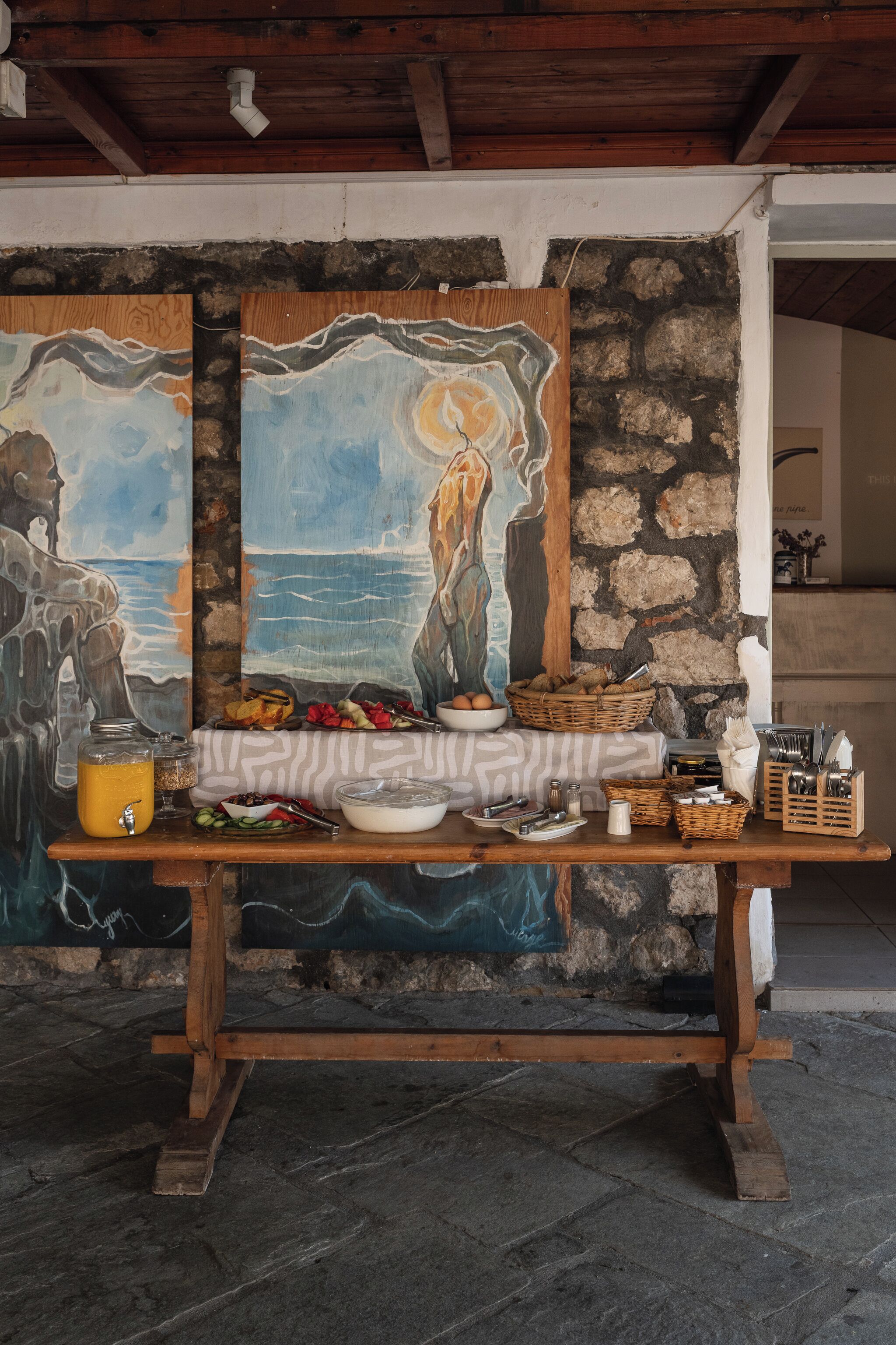 A rustic dining area with a stone wall backdrop, a wooden table set with various dishes, and a painting of a seascape.