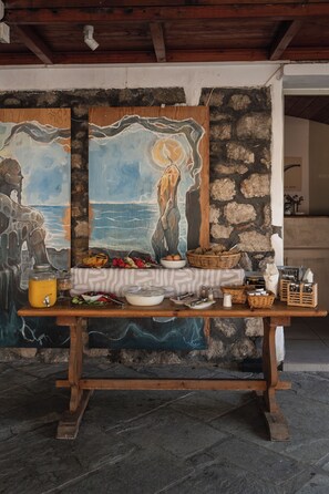 A rustic dining area with a stone wall backdrop, a wooden table set with various dishes, and a painting of a seascape. - Filanthi The Project (Pylos-Nestoras)