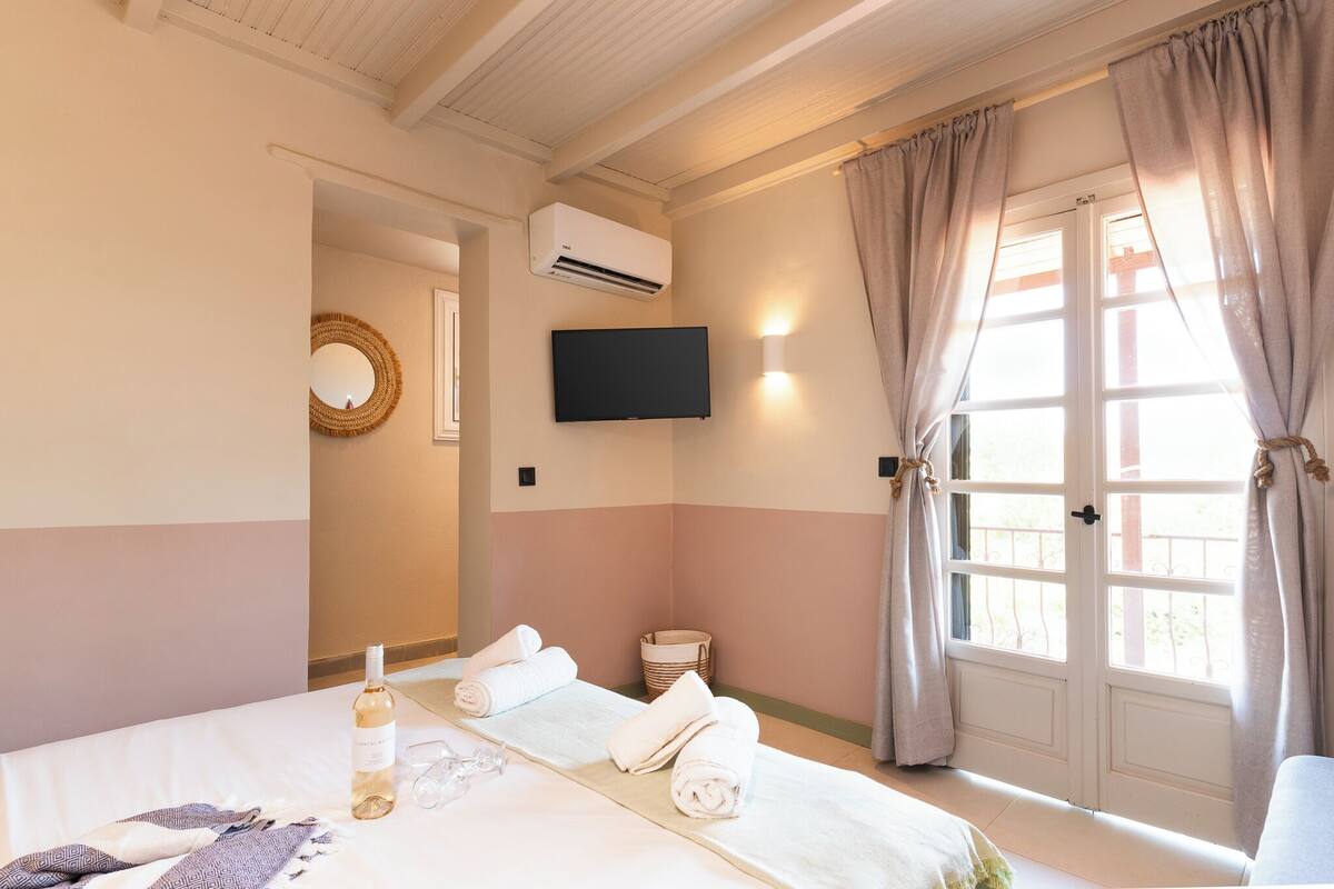 A hotel room with a bed, a flat-screen TV, an air conditioning unit, a window with curtains, and a small table with a bottle and glasses.
