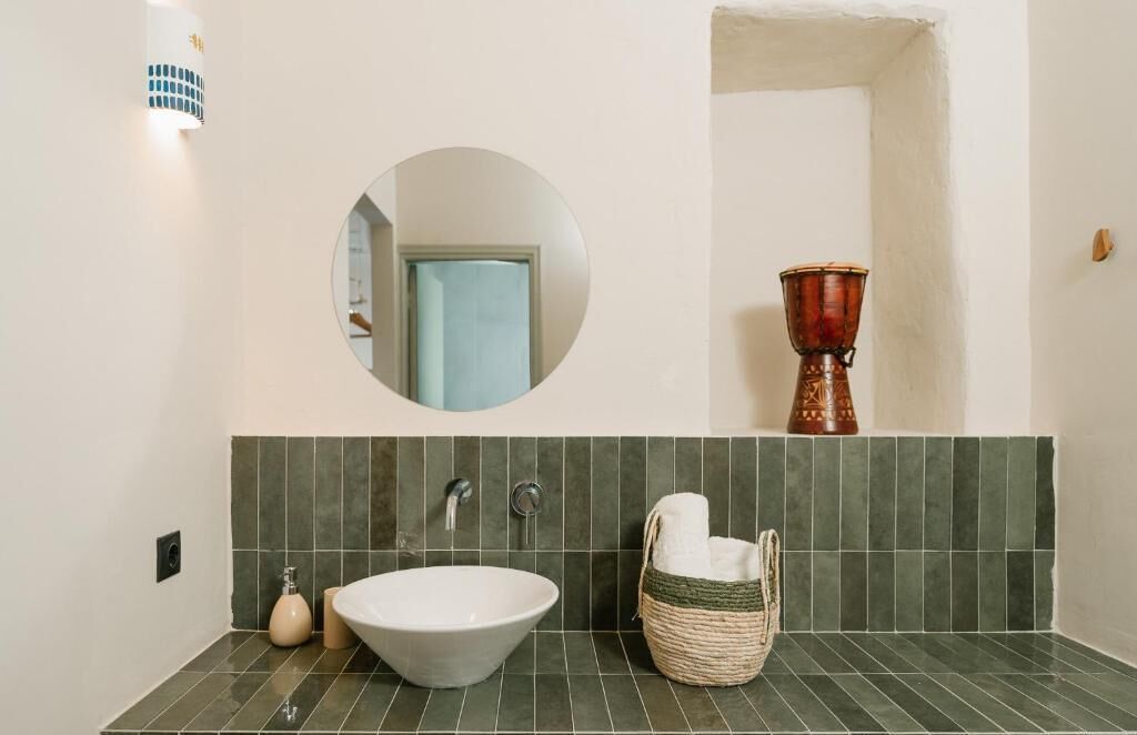 Une salle de bain avec un miroir rond, un lavabo blanc, un pare-feu en carreaux verts, un vase rouge et un panier en osier.