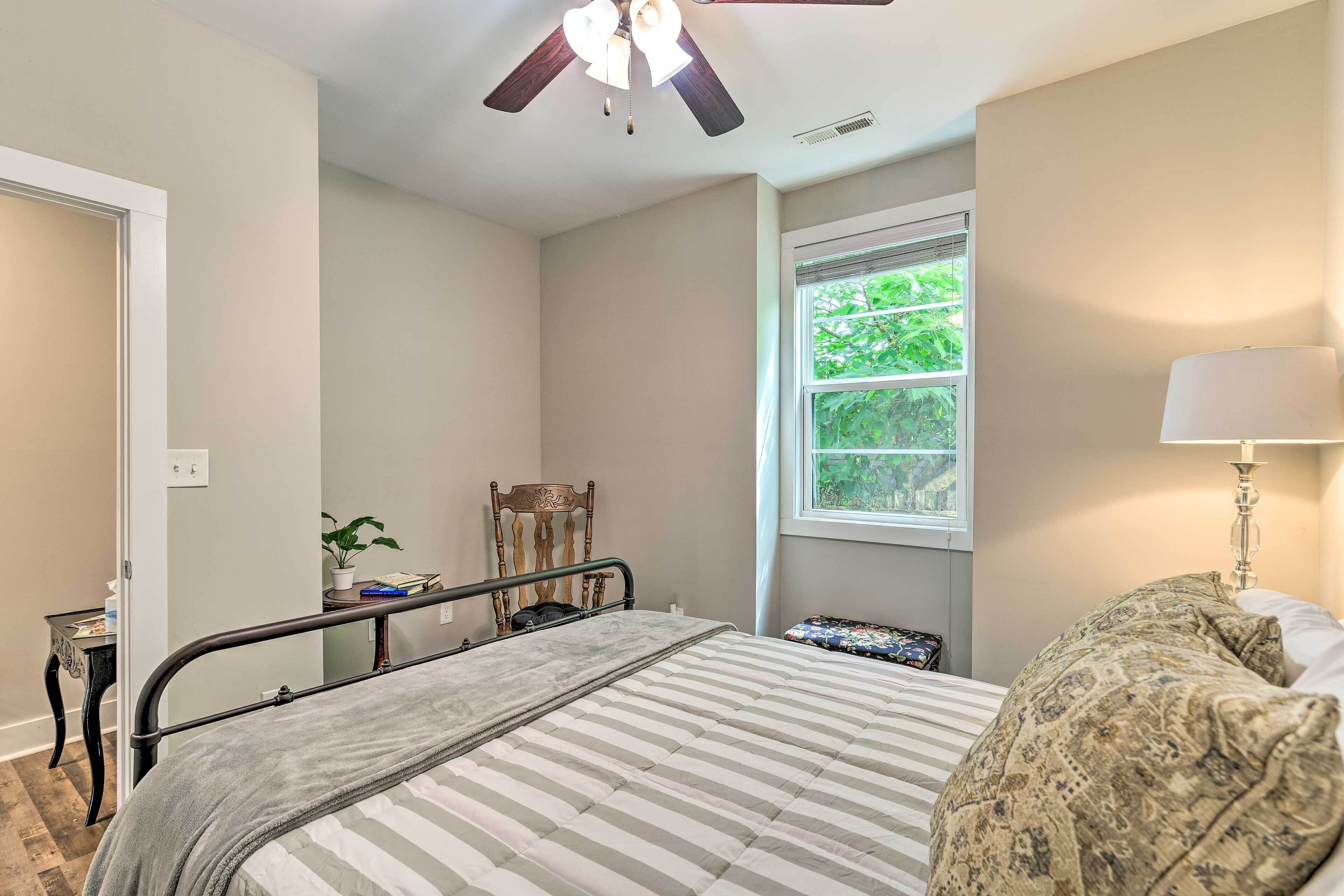 A bedroom with a bed, a ceiling fan, a window with a view of greenery, a bedside table, and a lamp.