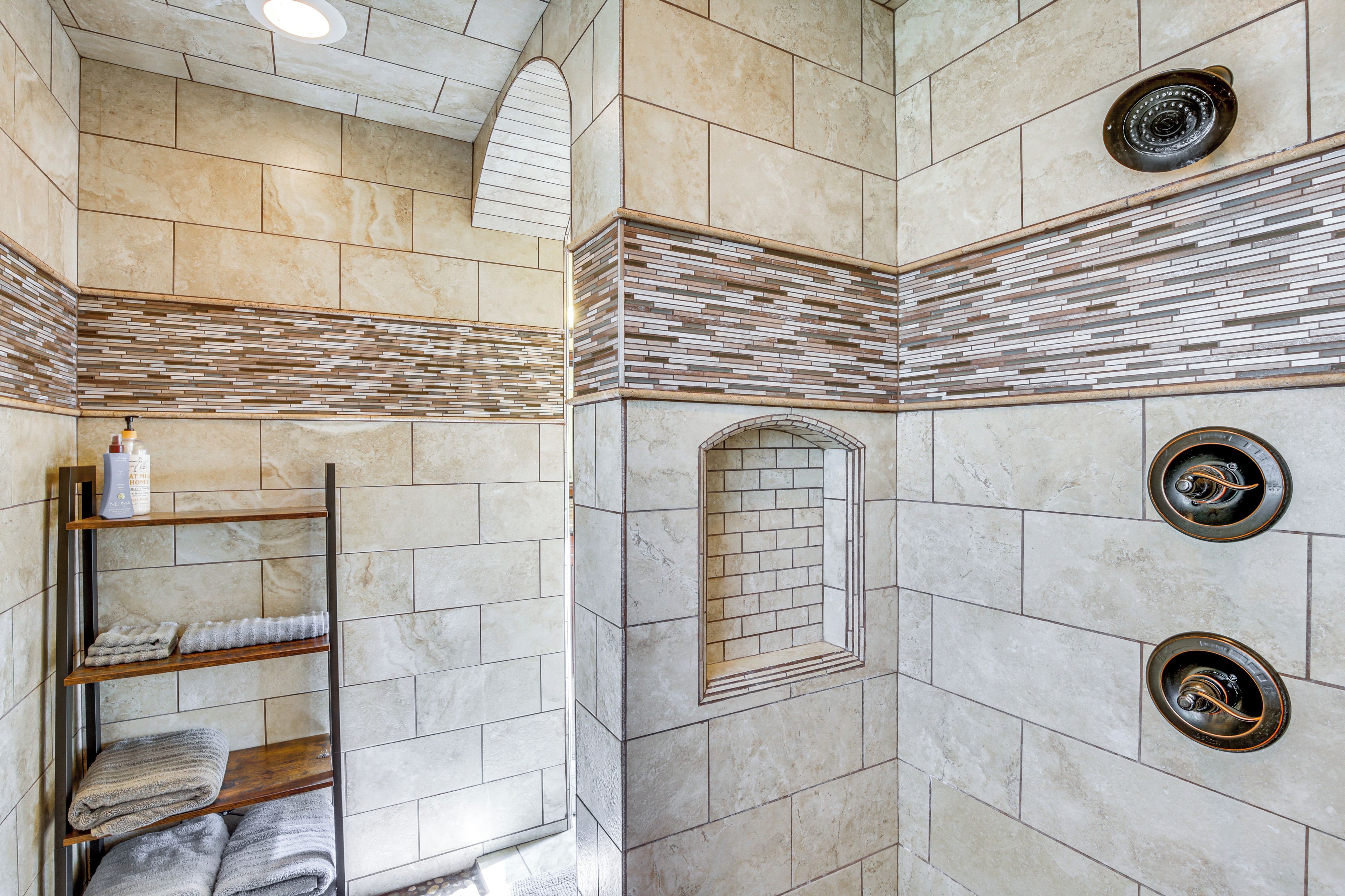 A bathroom with a tiled floor, a wooden shelf with towels, a shower with a circular drain, and a tiled wall with a brick pattern.