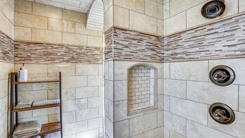 A bathroom with a tiled floor, a wooden shelf with towels, a shower with a circular drain, and a tiled wall with a brick pattern.