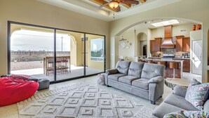 A spacious living area with a grey sofa, red bean bag, and patterned rug. A large sliding glass door leads to an outdoor patio with a table and chairs. The kitchen is visible in the background with wooden cabinets and a range hood. - Unique Las Cruces Home w/ Patio + Gas Grill! (Las Cruces)