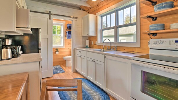 A compact kitchen with white cabinets, a stove, and a sink. There is a window providing natural light and a door leading to another room. - Cozy Cedarville Cottage - Walk to Lake Huron! (Cedarville)
