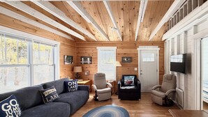 A cabin interior with a sofa, two armchairs, a TV, and a wooden ceiling. - Cozy Cedarville Cottage - Walk to Lake Huron! (Cedarville)