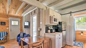 A cozy cabin interior with a wooden dining table, a kitchen area with a refrigerator and microwave, a blue bean bag chair, and a striped sofa. - Cozy Cedarville Cottage - Walk to Lake Huron! (Cedarville)