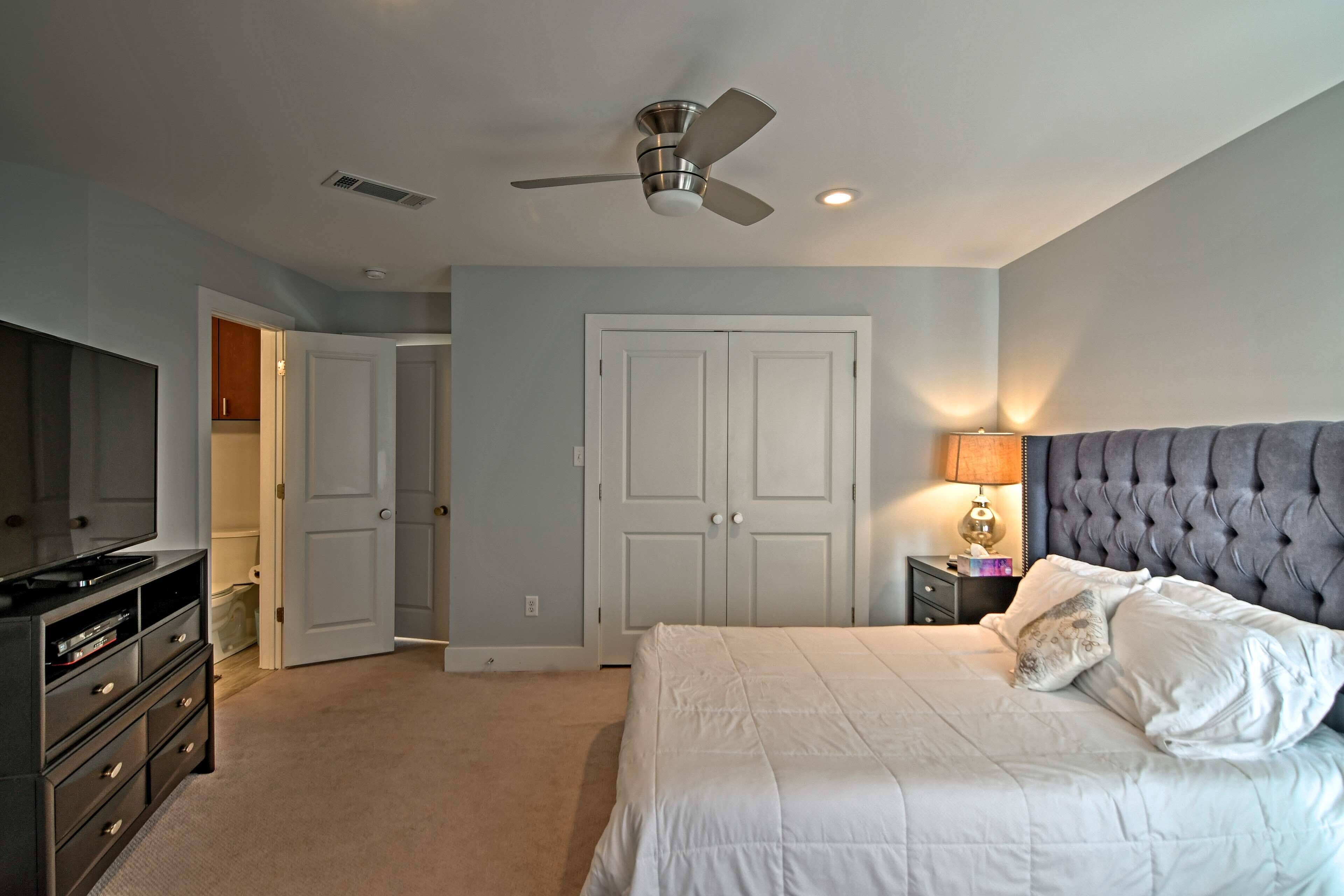 Un dormitorio con cama, ventilador de techo, televisión, cómoda y una puerta que da a otra habitación.