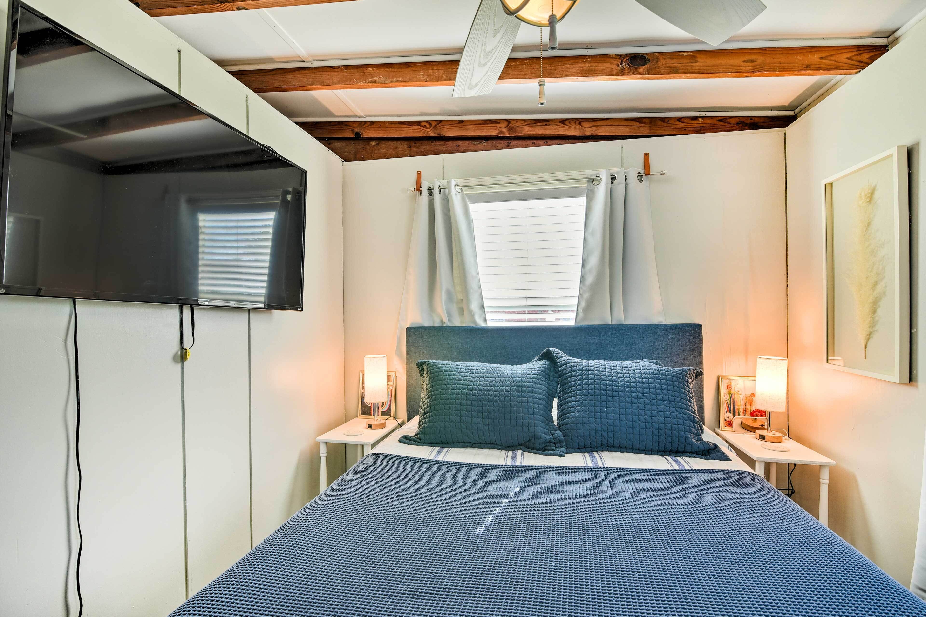 A bedroom with a bed, two bedside tables with lamps, a ceiling fan, a window with curtains, and a mounted TV.