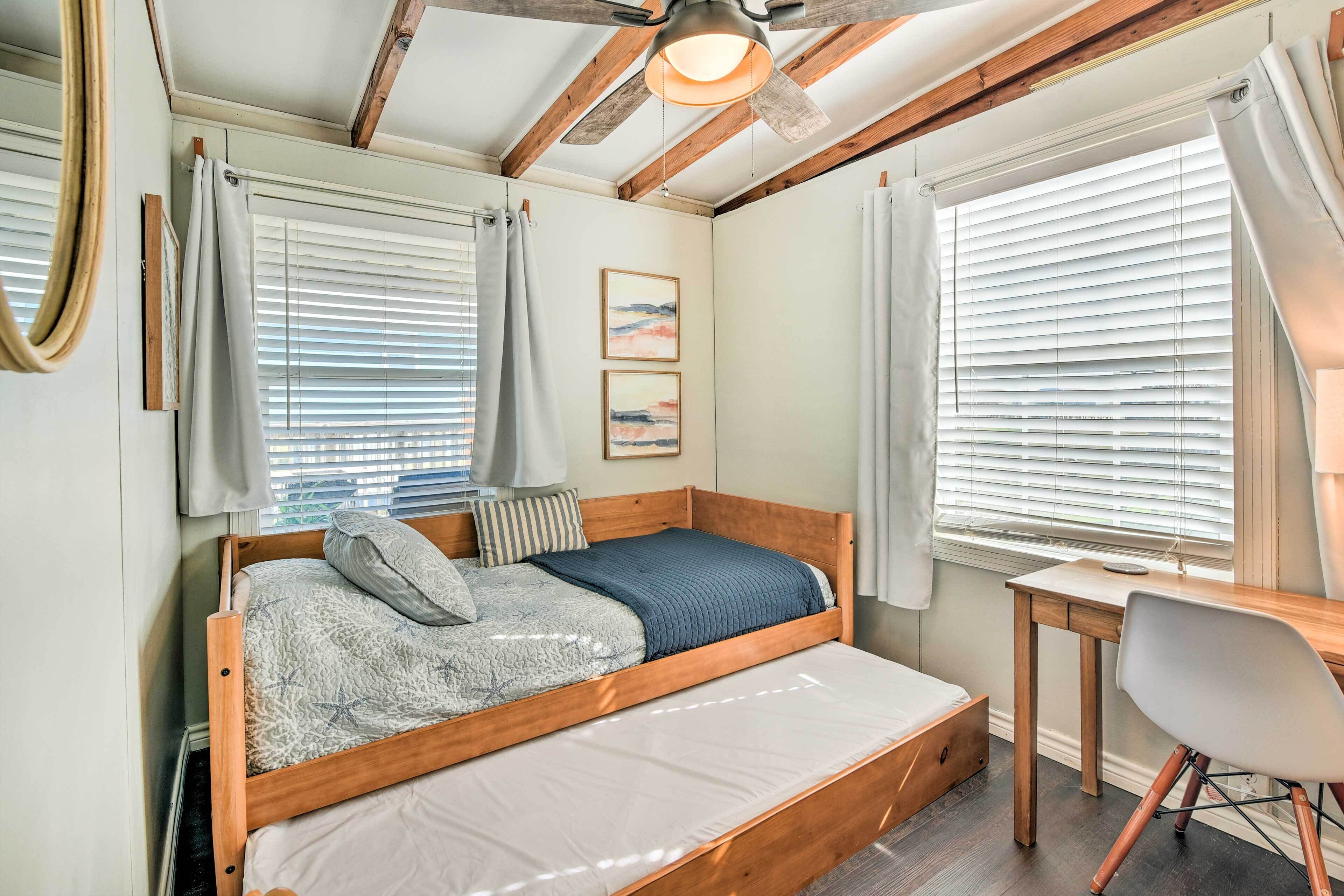 A bedroom with a wooden bed, a desk, and a chair. There are two windows with blinds and a ceiling fan.
